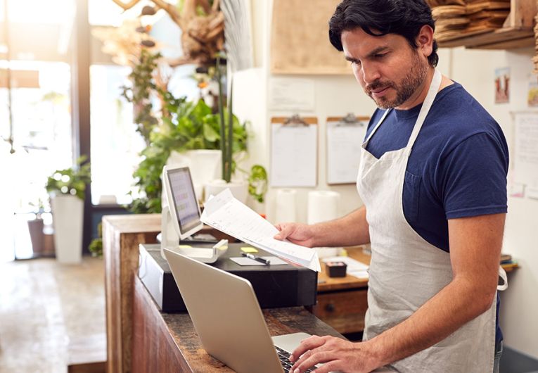 Male Sales Assistant Working On Laptop Behind Sales Desk Of Florists Store